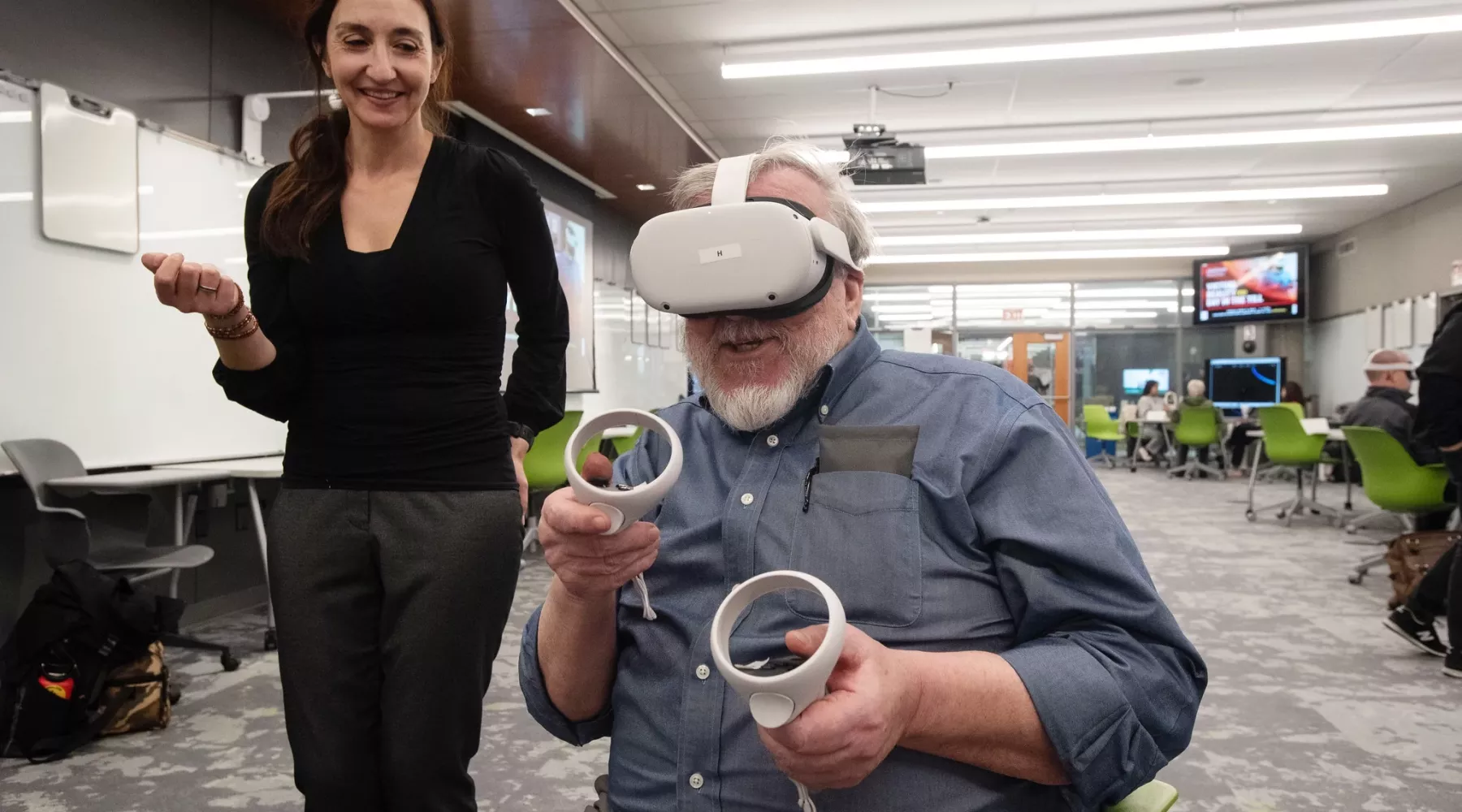 One person is seated using a VR set and hand controllers while someone else is standing looking at the VR participant.