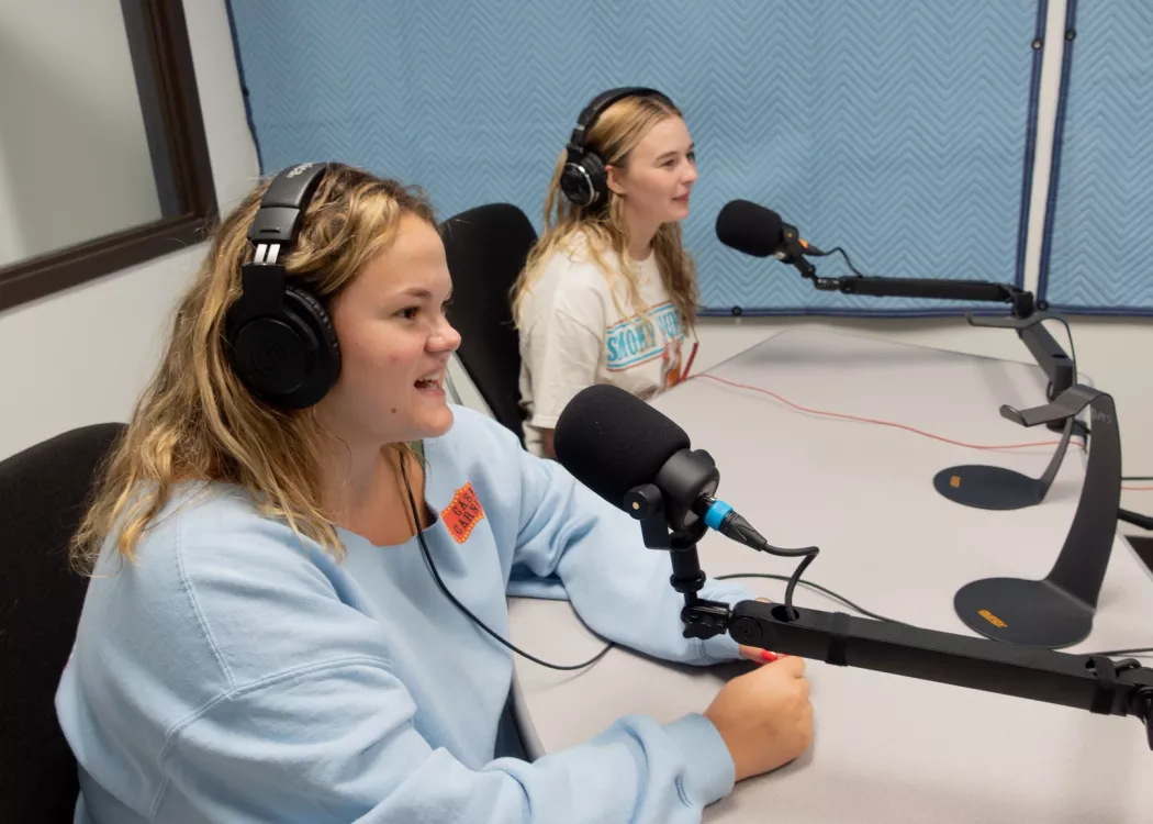 Two students sitting at a table with headphones, talking into podcast microphones.