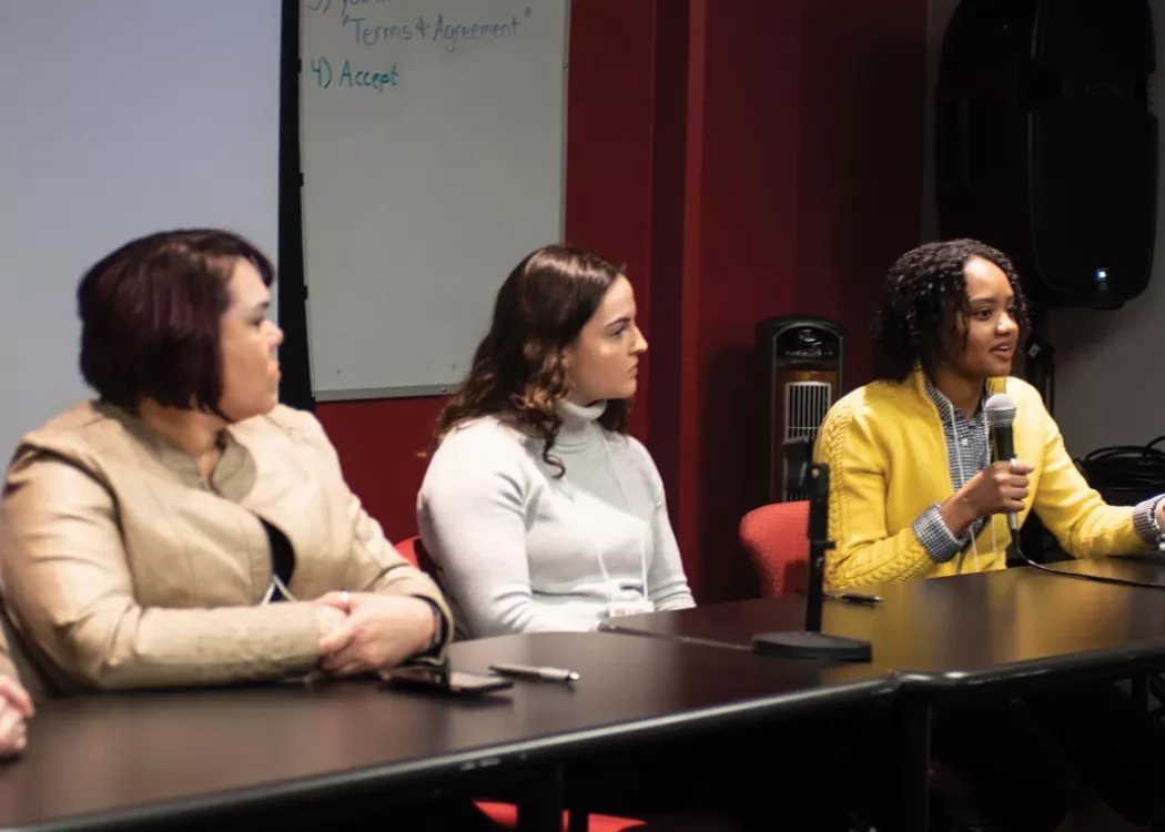 People sitting at a table with one person using microphone to speak to audience during a presentation